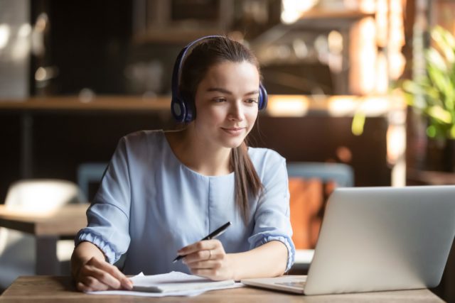 Focused woman wearing headphones using laptop, writing notes