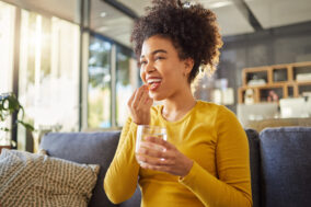 young-happy-mixed-race-woman-taking-medication-with-water-at-home-one-hispanic-female-with-a-curly-afro-taking-a-vitamin-for-good-health-while-sitting-on-the-couch-at-home-woman-drinking-a-supplemen